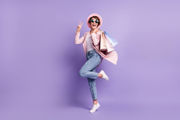 Full size photo of positive nice girl jump hold purchases show v-sign wear glasses coat jeans shoes isolated on purple background - 写真・画像