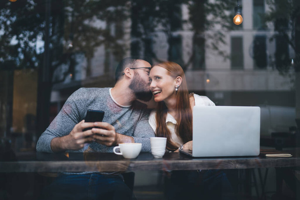 Junges Paar kuschelt am Cafétisch mit Tassen Heißgetränk und benutzt Laptop und Smartphone beim gemeinsamen Küssen - Foto, Bild