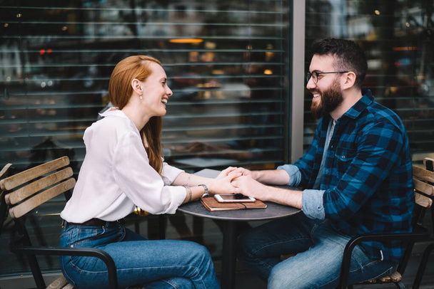 Side view of man in eyeglasses and woman in white shirt and jeans holding hands and laughing while sitting in cafe - Фото, изображение