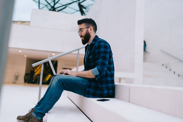 Side view of serious young bearded employee in checkered shirt and jeans and glasses using laptop while sitting on stairs in hall of contemporary business center - Фото, зображення