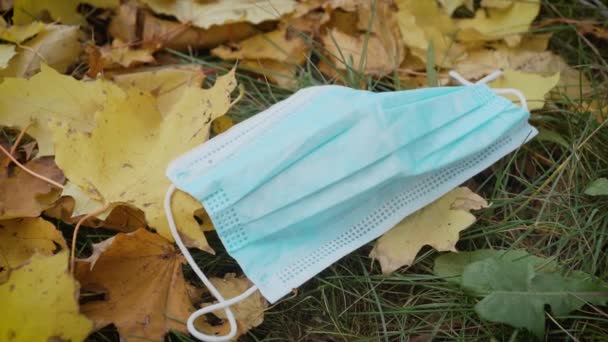 Gebruikt en uitgeworpen beschermend medisch masker voor coronavirus preventie liggend op gras met geel gevallen bladeren in stadspark - Video