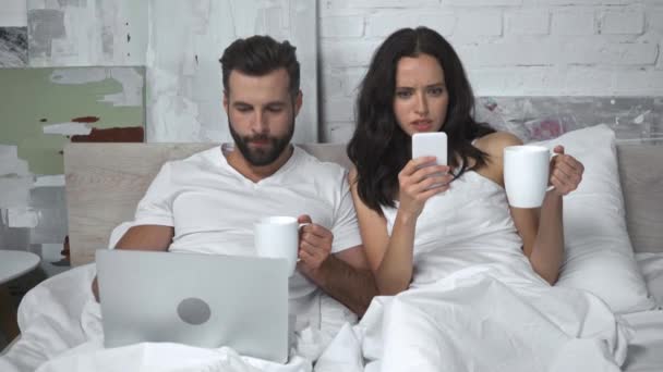 bärtiger Mann benutzt Laptop und trinkt Tee in der Nähe von Frau mit Smartphone - Filmmaterial, Video