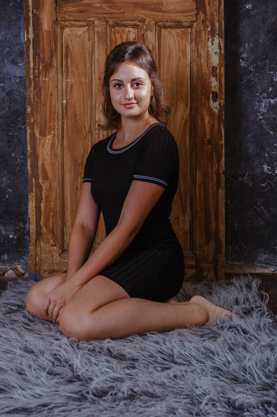 Beautiful young woman in black dress sitting on fur skin in front of vintage door - Photo, Image