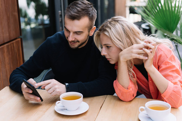Focused couple in casual wear browsing smartphone and attentively looking at screen together while drinking tea in street cafe at daytime - Foto, afbeelding