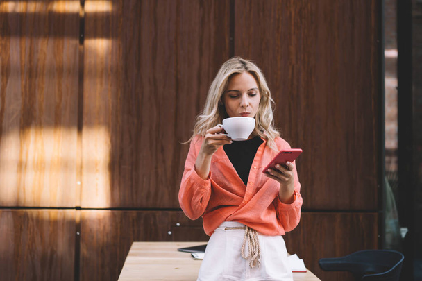Spokojna kobieta w ubraniach casual przeglądania telefonu komórkowego podczas picia filiżanki gorącego napoju i stojąc przy stole w kreatywnej przestrzeni roboczej - Zdjęcie, obraz