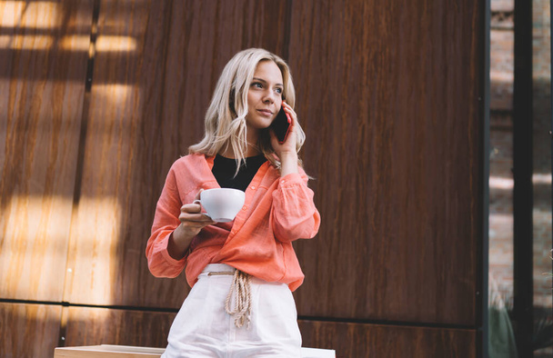 Low angle of calm young female customer in boho outfit drinking coffee and talking on smartphone looking away while standing near table in creative cafeteria with wooden interior at daylight - Foto, immagini