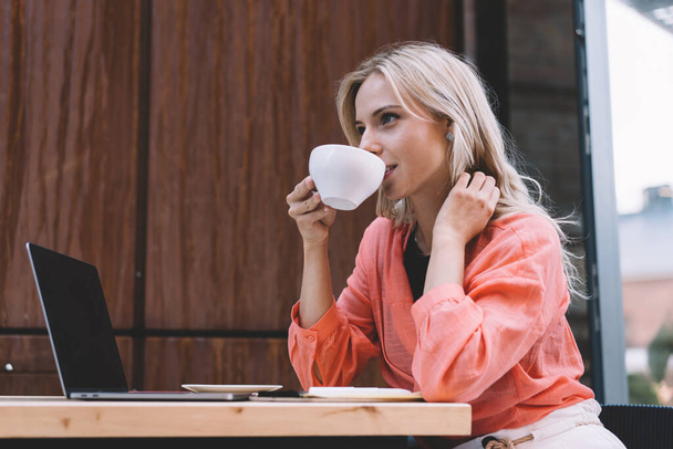 Вид збоку позитивного фрілансера жінки в рожевій випадковій блузці, що п'є чашку кави, сидячи за столом з ноутбуком під час перерви в роботі в творчій кафетерії
 - Фото, зображення