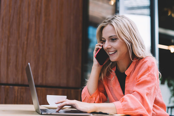 Smiling female freelancer in casual blouse using laptop during phone call while sitting at table with cup of coffee working remotely in modern cafeteria - Fotó, kép