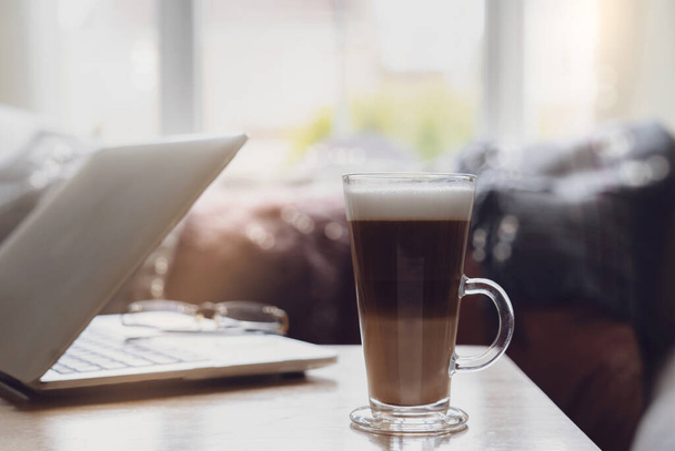 Házi készítésű szemüveg latte kávébab és laptop dohányzóasztalon a munkahelyen ablak mellett reggeli fény vagy hangulatos jelenet pihentető délutáni szünet kávé és notebook a nappaliban - Fotó, kép