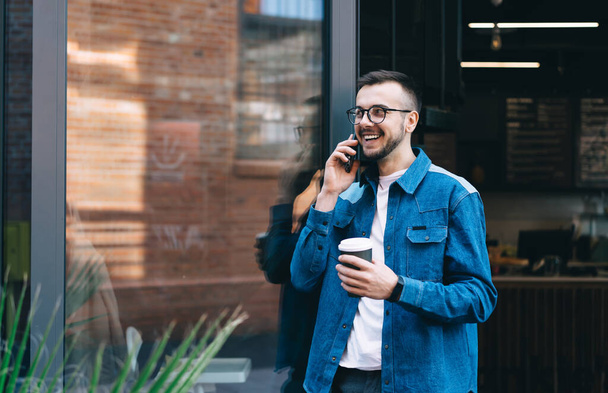 Happy young man in casual wear and eyeglasses answering phone call while holding takeaway coffee and leaning on door of modern cafe - Foto, Bild