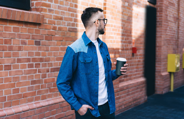 Young man in jean jacket and eyeglasses holding hand in pocket and looking away while drinking takeaway tea against brick wall - Φωτογραφία, εικόνα