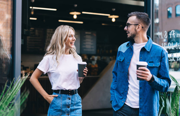 Cheerful casually dressed couple with paper cups of coffee smiling and looking at each other after coming out of cafe - Foto, afbeelding