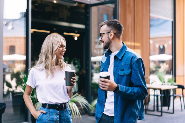 Positive young couple with takeaway coffee enjoying conversation while toothy smiling and looking at each other near modern street cafe - Foto, Bild