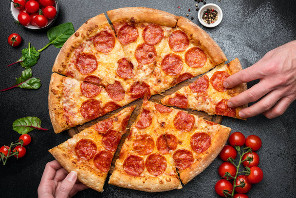 Pepperoni pizza on black concrete background. Hand picking pepperoni pizza slice. Top view - Foto, Bild