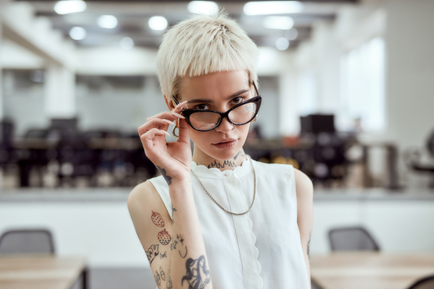 Young and succesful. Portrait of confident blonde tattooed businesswoman adjusting her eyeglasses and looking at camera while standing in the modern working space - Фото, изображение