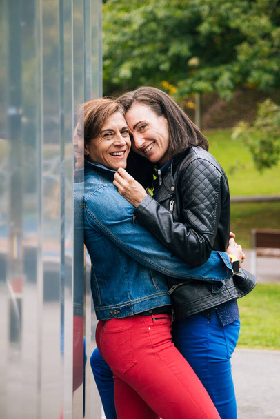 Happy middle aged lesbian couple hugging against a metallic wall and looking to camera - Foto, Imagen