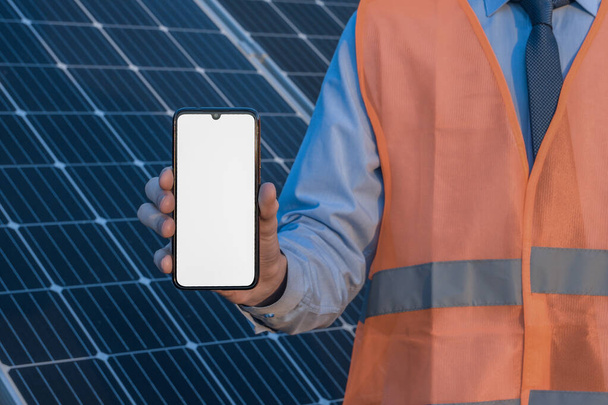 Ingénieur, homme tenant un téléphone portable, smartphone sur fond de panneaux solaires. Écran blanc, endroit pour l'image, texte, application. - Photo, image