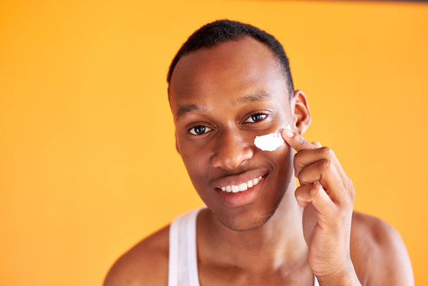 young african-american guy applying face cream under his eyes on yellow background - Φωτογραφία, εικόνα