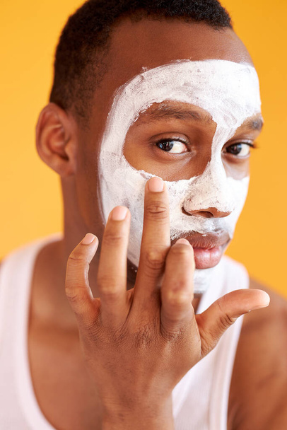 young african-american guy applying face cream under his eyes on yellow background - Foto, Imagem