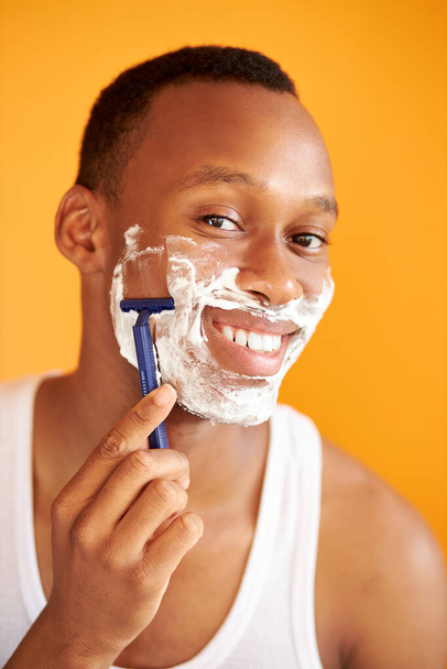 portrait of african male shaving his face with foam and razor isolated on orange background - Zdjęcie, obraz