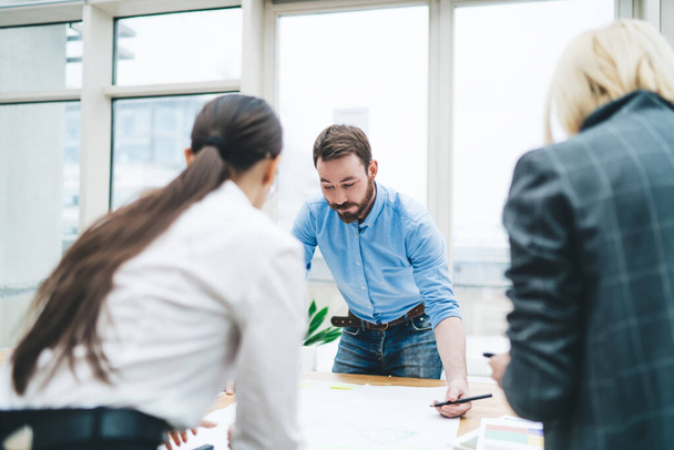 Concentrated bearded male in blue shirt jeans focusing on working with project on table standing opposite faceless female colleagues in office - Foto, Imagen
