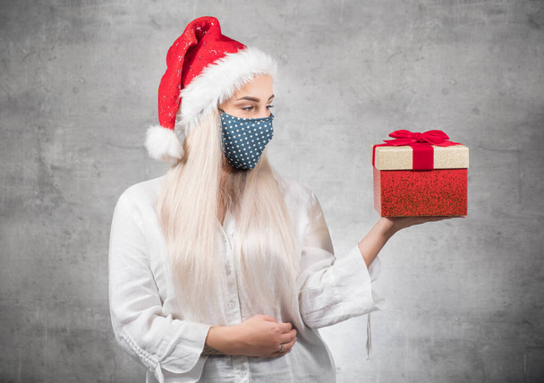 Santa woman shows christmas gift isolated on gray background. Happy young girl wearing red santa hat and holding present box. A Corona - Covid 19 Christmas concept. Studio shot - Φωτογραφία, εικόνα