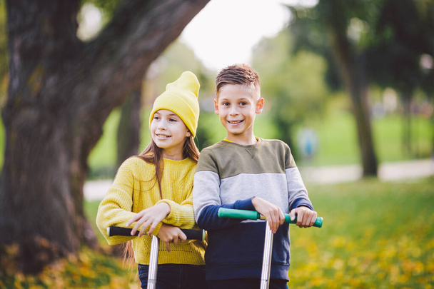 Двое детей кавказский мальчик и девочка близнецы позируют со скутерами в осеннем парке. Счастливые дети катаются на скутерах в общественном парке. Детский спорт. Занятия на свежем воздухе. Счастливого детства - Фото, изображение