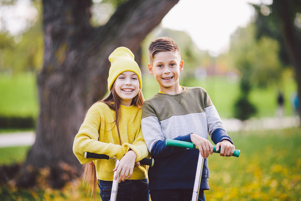 白人の子供たち双子の男の子と女の子の子供たちは、キックスクーターに乗って秋の公園で晴れた天気の中で一緒にポーズをとります。公園でキックスクーターに乗る幸せな子供たち。キッズスポーツ. - 写真・画像