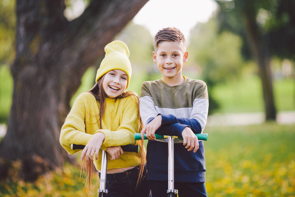 Tweeling 10 jaar oud in heldere kleren tijd actief samen doorbrengen in de herfst park rijden kick scooters. Gelukkige kinderen. Eco-vervoer. Kinderen poseren in het herfstpark. Broer en zus pre tiens portret. - Foto, afbeelding