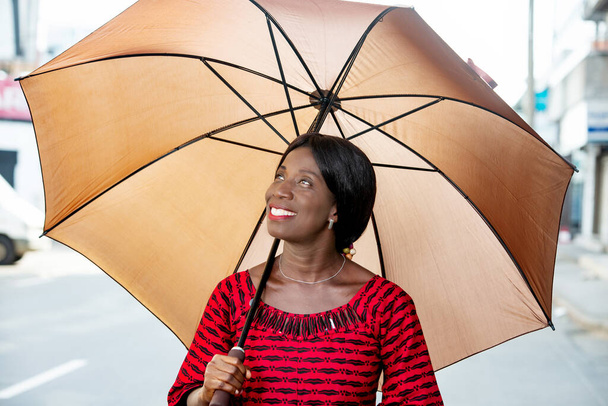 beautiful african adult businesswoman dressed in red loincloth holding umbrella in hand and looking up smiling in the street - Фото, зображення