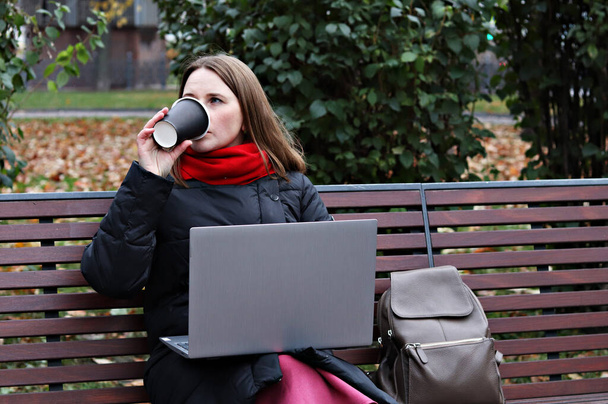 giovane donna si siede su una panchina del parco, beve caffè e lavora in un computer per strada. - Foto, immagini