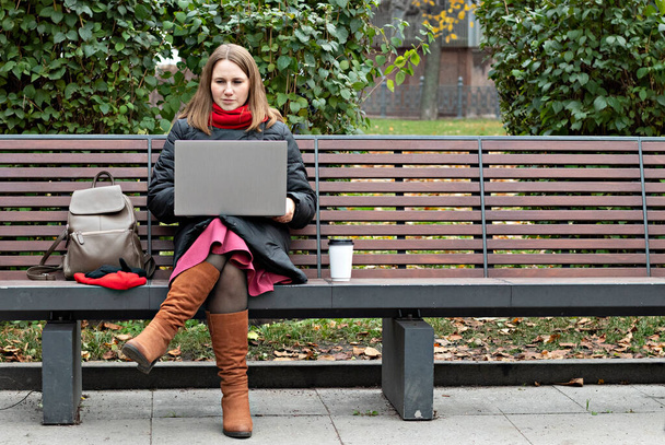 mujer joven se sienta en un banco del parque y trabaja en una computadora en la calle. - Foto, Imagen