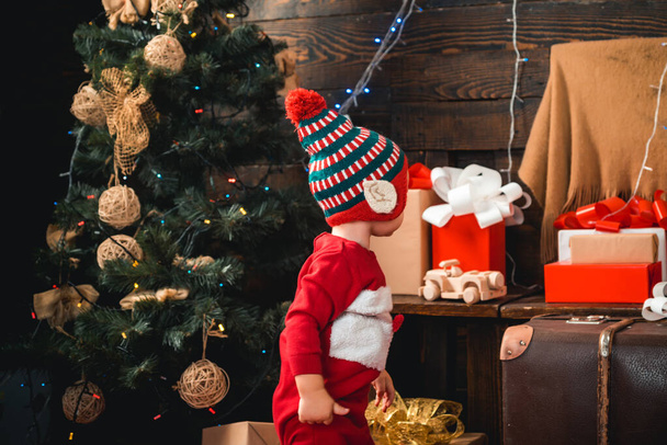 Babies. Happy small kids in santa hat with present have a christmas. Portrait kid with gift on wooden background. Winter kids. Portrait of happy child looking at decorative toy ball by Christmas tree. - Photo, image