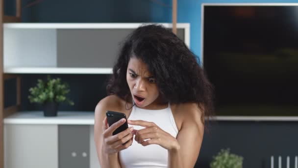 Mulher afro-americana chocada lendo notícias terríveis em seu smartphone em casa apartamento fundo. Desagradavelmente surpreendido jovem fêmea reagindo mensagem em seu telefone celular. - Filmagem, Vídeo