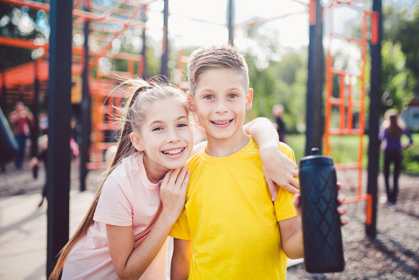 Caucasian children brother and sister stand in embrace at outdoor sports ground with smile and flask with water after training. Twins boy and girl athlete posing looking at camera during workout break - Photo, image