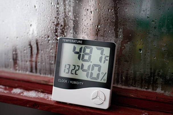 ガラス上の結露の背景に電子時計、カレンダー、温度計、および湿度計、高湿度。研究室のデジタル湿度計は、薬局方の温度を示しています - 写真・画像