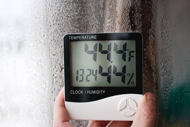 Electronic clock, calendar, thermometer, and hygrometer, against the background of condensation on glass, high humidity. Digital hygrometer in the laboratory shows the temperature of the pharyngeite - Photo, Image