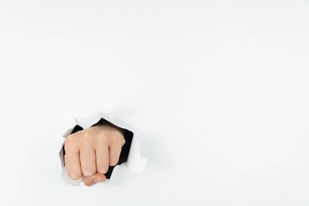 Die zu einer Faust geballte Hand einer Frau durchbohrt die weiße Papierwand. Die Hand geht durch das Papier. Aggressionskonzept. Raum für Text - Foto, Bild