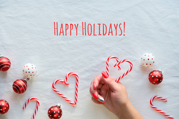 Christmas background, flat lay, top view on white textile background. Female hands holding heart shapes made from candy canes. Red and white trinkets with dots. - Foto, afbeelding