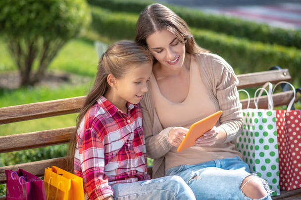 Smiling mom and daughter sitting on park bench, checking tablet - Фото, изображение