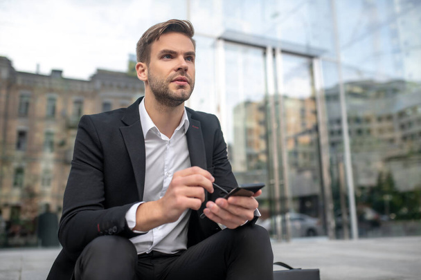 A businessman writing a message on a smartphone and looking thoughtful - Photo, Image
