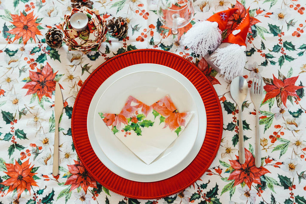 Christmas table decoration with floral tablecloth, decorative elements, glass of wine, white plates on a red trivet and aluminum cutlery. Christmas concept. Flat lay. - Фото, изображение