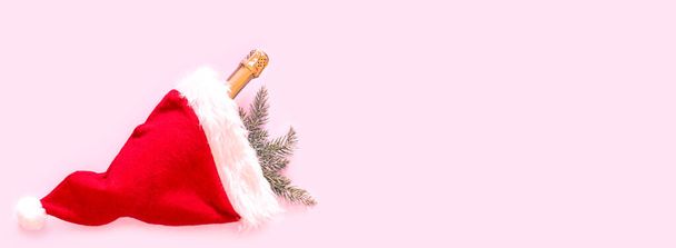 Rode Kerstman hoed, sparren takken en fles champagne op roze achtergrond. Kerstmis en Nieuwjaar concept. Platte lay, bovenaanzicht, kopieerruimte. Banner - Foto, afbeelding