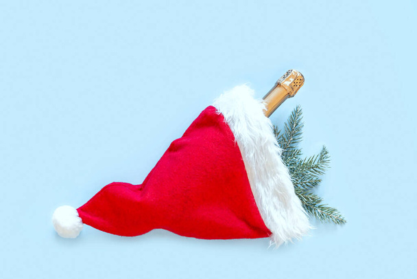 Κόκκινο καπέλο Σάντα, κλαδιά ερυθρελάτης και σαμπάνια μπουκάλι σε μπλε φόντο. Χριστούγεννα και Πρωτοχρονιά έννοια. Επίπεδο lay, πάνω όψη, αντιγραφή χώρου. - Φωτογραφία, εικόνα