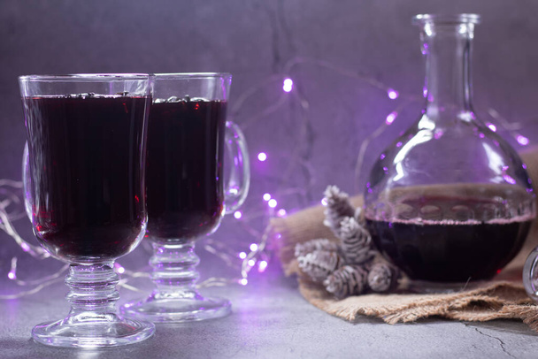 Two transparent cups of mulled wine with cinnamon and cardamom inside and a decanter on a gray background and purple lights. - Photo, Image