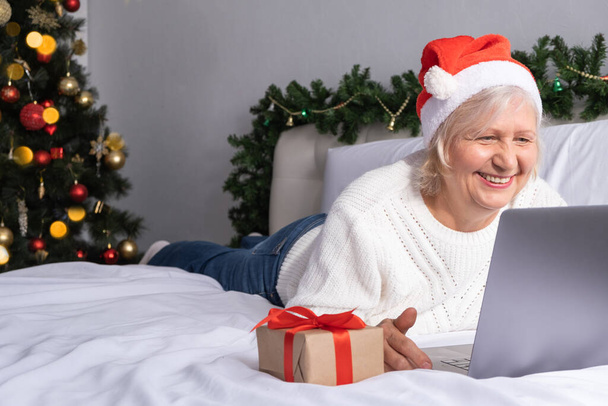 Natale saluti online. felice donne anziane a Santa cappello a casa a letto con il computer portatile. - Foto, immagini