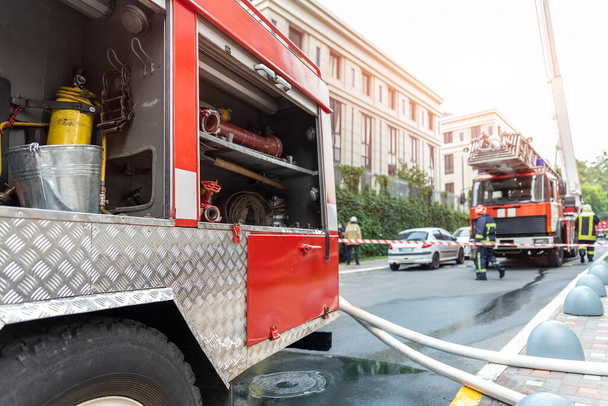 Πυροσβέστες φορώντας στολή στέκεται πίσω από ταινία φωτιά και πολλά πυροσβεστικά οχήματα με σκάλα κατά λάθος σε πύργο highrise κατοικιών ή κτίριο γραφείων στο κέντρο της πόλης. Επείγουσα διάσωση σε περίπτωση καταστροφής - Φωτογραφία, εικόνα