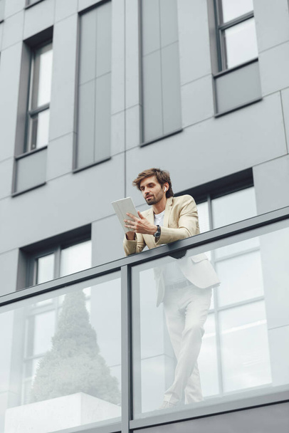 Konzentrierter junger Geschäftsmann im schicken Anzug, der auf dem Balkon des eigenen Büros steht und ein modernes Tablet in der Hand hält. Bärtiger Mann arbeitet im Freien an digitalem Gerät. - Foto, Bild