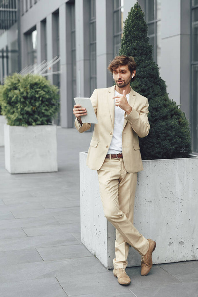 Full length portret van bebaarde jonge zakenman in beige pak met videogesprek op digitale tablet buiten. Zelfverzekerde man in draadloze oortelefoons werkt op afstand. - Foto, afbeelding
