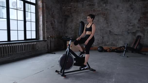 Young Athletic Indian Woman taking a Break from Training, Sitting on a Bike Dressed in Sportswear Black Top and Leggings, Is in the Gym - Záběry, video